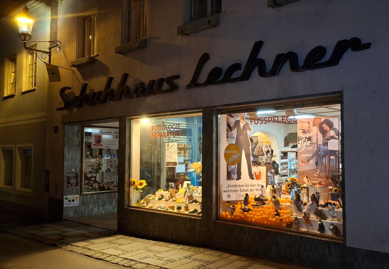 Das Schuhhaus Lechne Geschäft mit beleuchtetem Schild. Das Schaufenster zeigt einen Dummy, Schuhe und Sonnenblumen. Der Name des Geschäfts und "Podologie Reparatur" sind im Fenster.