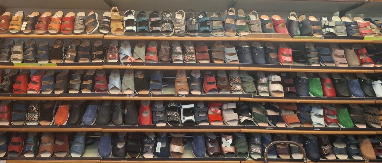 Ein Geschäft zeigt mehrere Reihen von Sandalen in verschiedenen Farben und Stilen auf Holzregalen. Die Sandalen haben verschiedene Designs, Schnallen und Riemen.