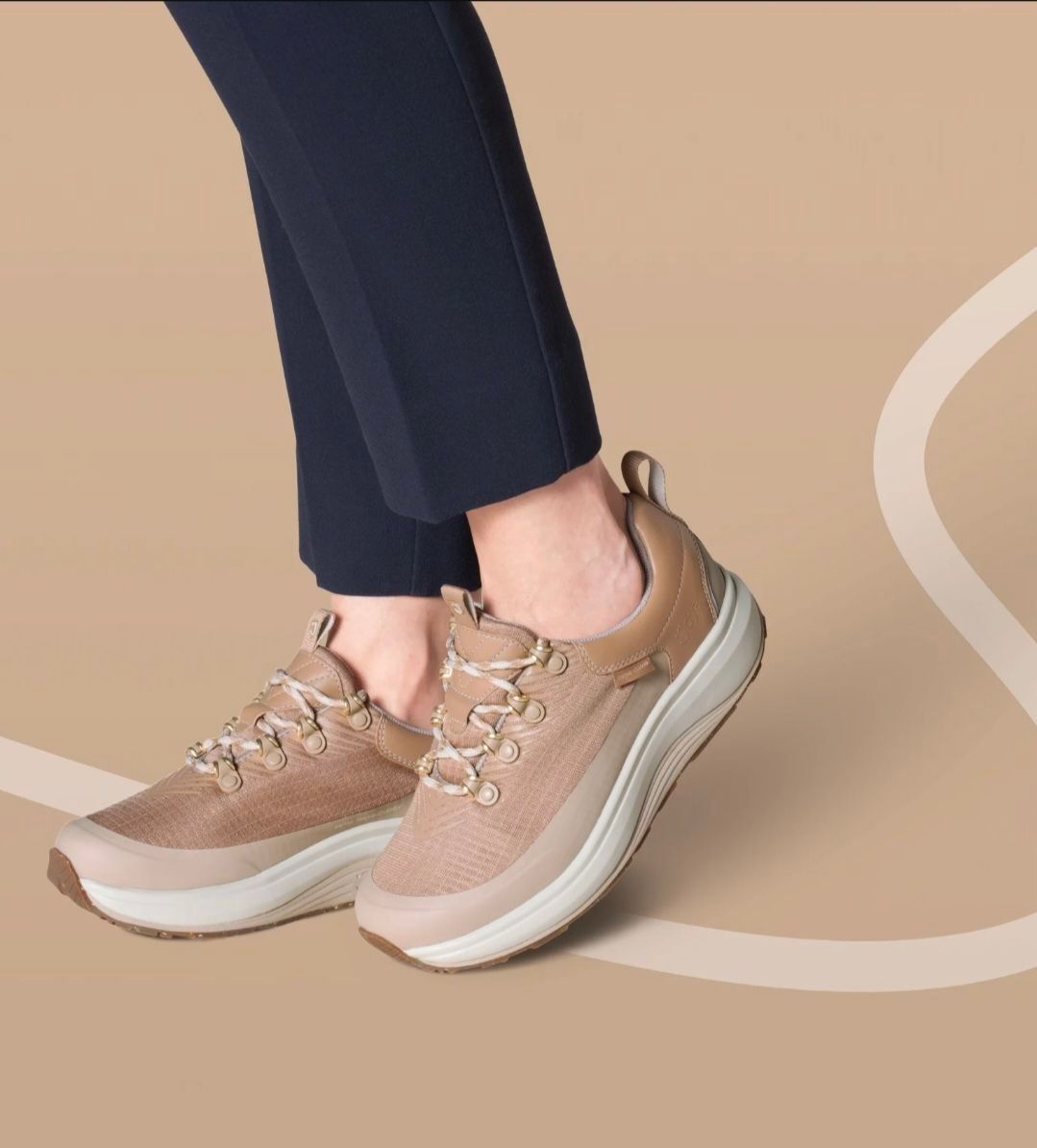 Eine Person, die blaue Hosen und ein Paar beigefarbene und weiße Turnschuhe mit goldenen Schnürsenkeln auf einem beigefarbenen Hintergrund trägt.