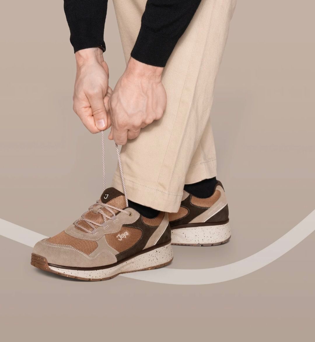 Eine Person in khakifarbener Hose und schwarzem Langarmshirt bindet eine Schnürsenkel an einem braunen und beigen Sneaker.