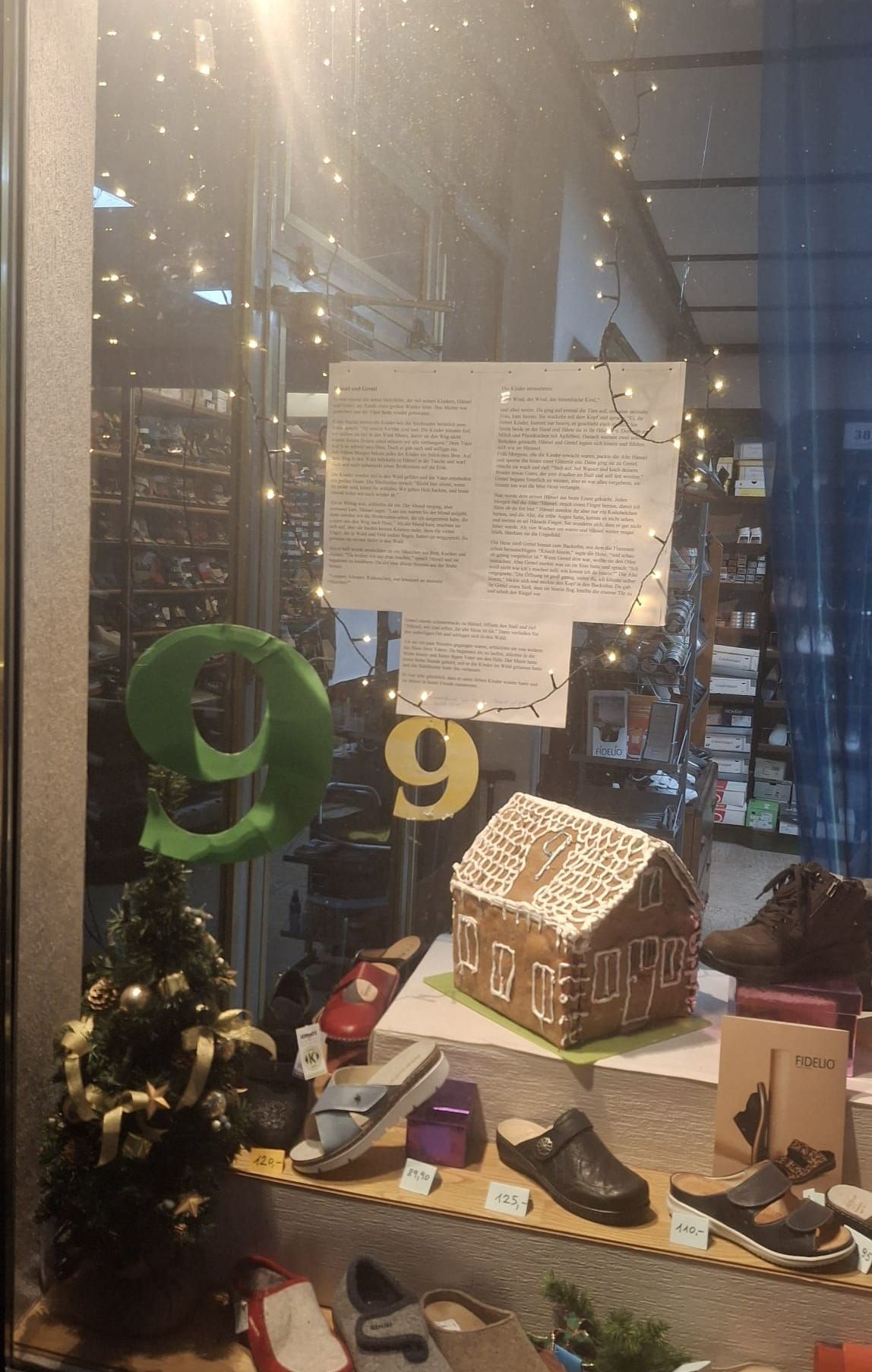 Ein Schaufenster zeigt ein Lebkuchenhaus und Schuhe. Ein Papier mit Text hängt darüber und eine grüne Zahl 9 ist auf dem Glas. Ein Weihnachtsbaum und weitere Schuhe sind im Vordergrund.