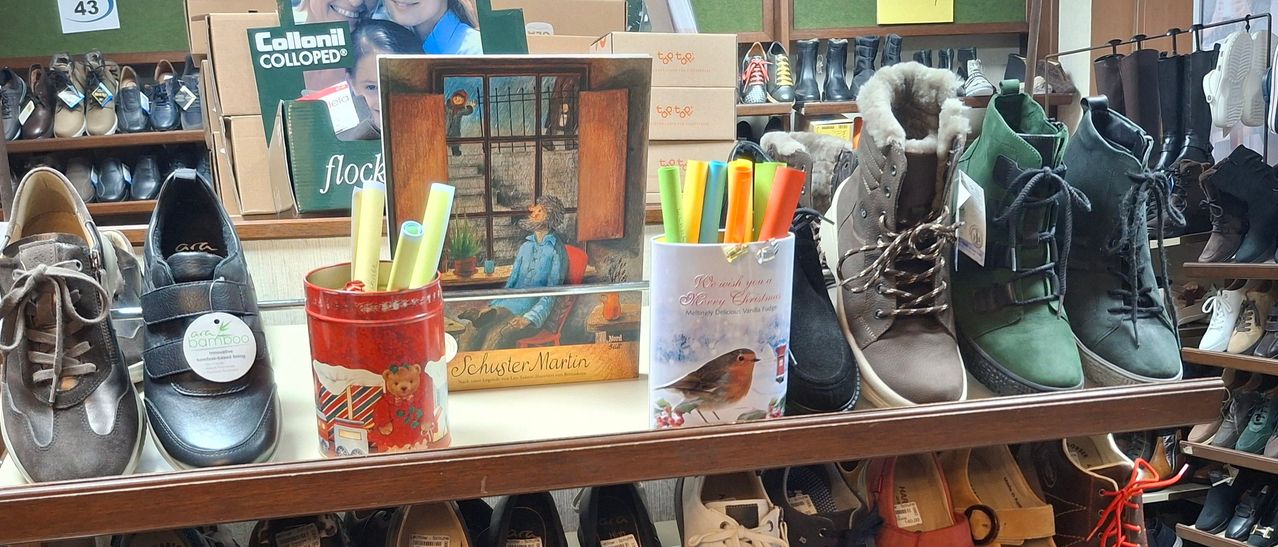 Ein Regal hält eine Schachtel Martin Schuster Bücher und einen Behälter mit farbigen Markern. Hinter der Schachtel ist ein Mädchen in einem blauen Shirt zu sehen. Schuhe und Stiefel sind im Hintergrund.