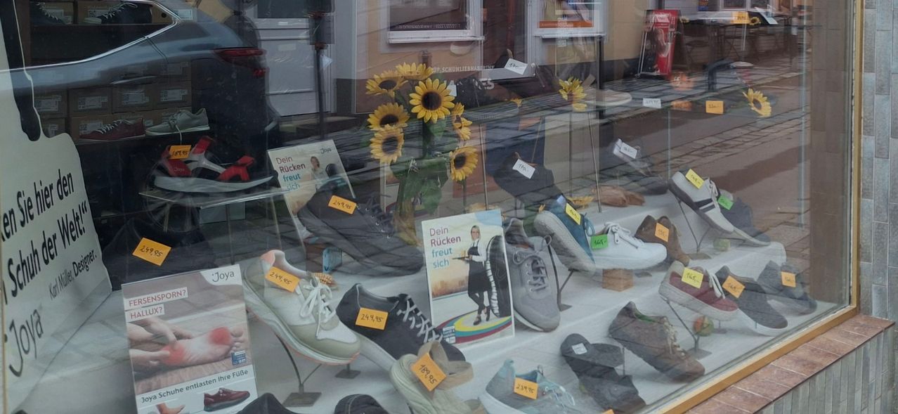 Ein Schaufenster zeigt mehrere Paar Schuhe mit Preisschildern, einen Strauß Sonnenblumen und ein Poster mit einer Frau in einer Kochuniform.