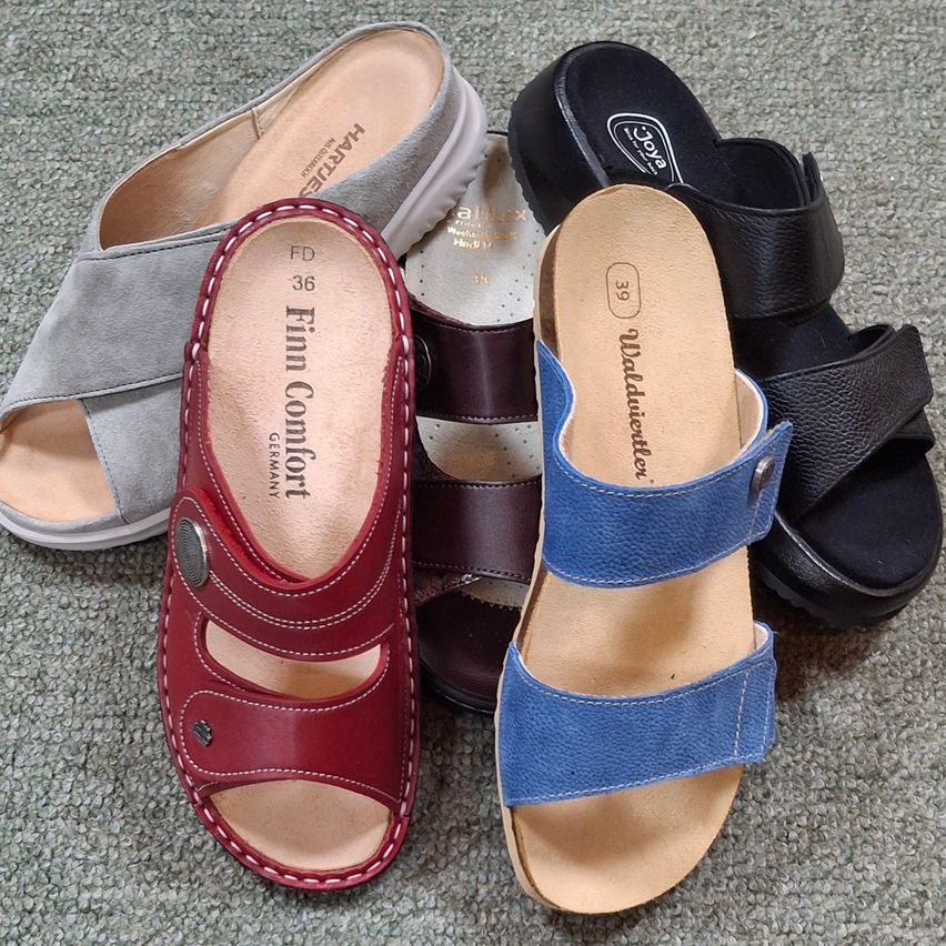 Fünf Paar Sandalen liegen auf einem grauen Teppich. Sie umfassen ein rotes, ein blaues, ein schwarzes, ein braunes und ein graues Paar. Jedes Paar hat ein anderes Design und Material.