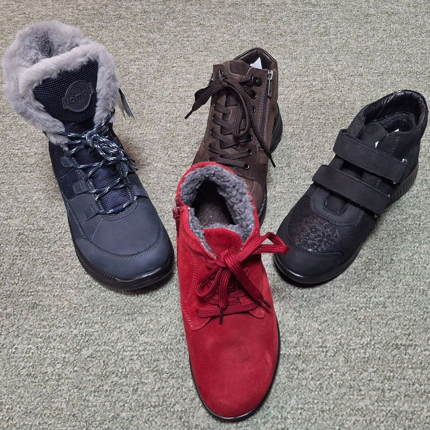 Drei Paar Winterstiefel auf einem Teppichboden. Der linke Stiefel ist marineblau mit Pelzkragen, der mittlere ist rot aus Wildleder mit einem Pelzkragen und der rechte Stiefel ist schwarz mit einem Riemen.