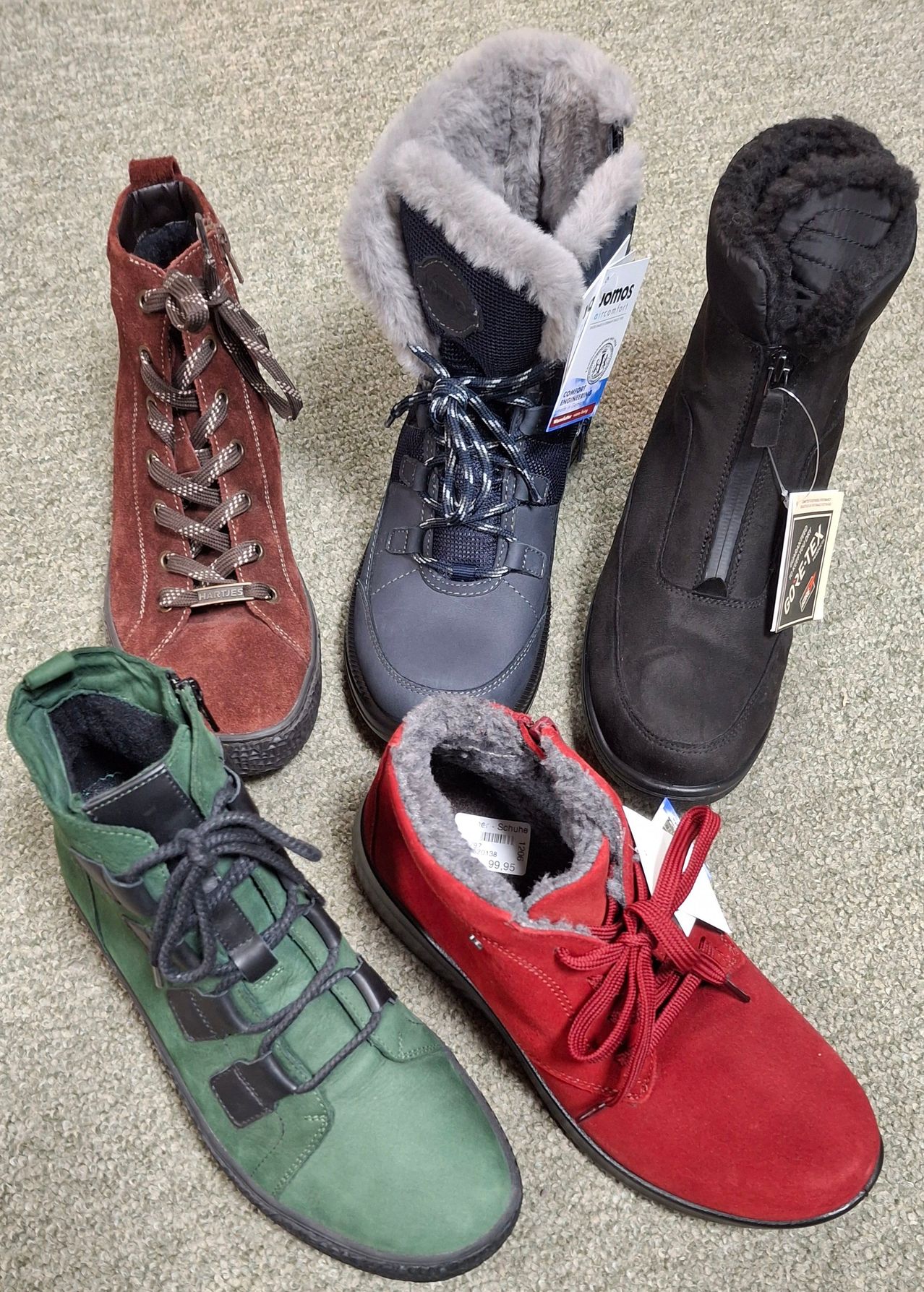 Fünf Paar Stiefel in verschiedenen Farben und Stilen sind auf einem Teppichboden ausgelegt. Sie haben Schnürsenkel, Reißverschlüsse und Fellfutter. Jedes Paar hat einen Preisschild.