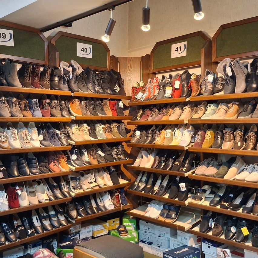 Ein Geschäft mit Regalen voller verschiedener Schuhe, darunter Stiefel, Sneaker und Sandalen, sortiert nach Größe und mit Preisschildern. Die Regale sind gut beleuchtet und organisiert, mit Kartons am Boden.