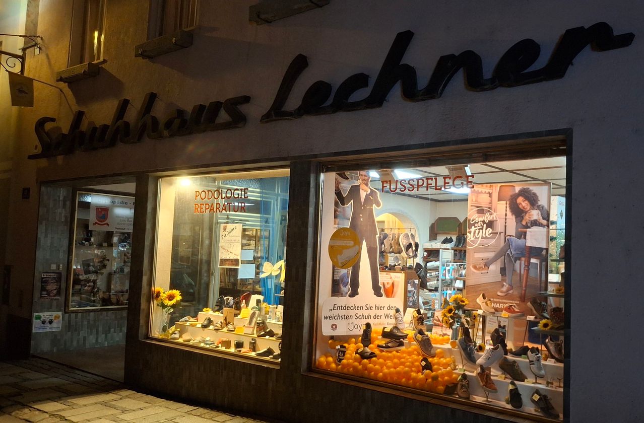 Ein Laden mit einem Schild, das 'Haus Lechner' steht. Im Schaufenster sind verschiedene Schuhe und ein Poster eines Mannes in Anzug mit dem Text 'Podologie Reparatur Fusspflege' zu sehen. Im Fenster sind Schuhe und Sonnenblumen zu sehen.