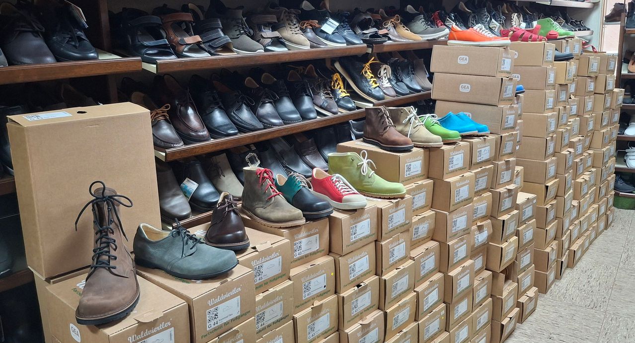 Ein Schuhgeschäft zeigt zahlreiche Paar Schuhe auf Regalen und gestapelten Kartons, einschließlich Stiefel, Sneaker und Dress-Schuhe in verschiedenen Farben und Stilen.