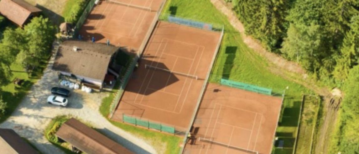 Luftaufnahme mehrerer Tennisplätze, umgeben von Bäumen und Gebäuden. Zwei Autos geparkt vorn.
