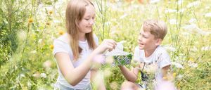 Ein Mädchen und ein Junge sitzen in einem Feld und lächeln. Das Mädchen hält eine Taschentuchbox. Der Junge schaut sie an.