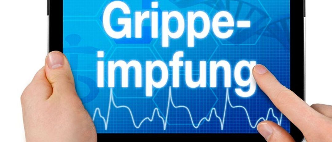 Eine Person hält ein Tablet mit einem blauen Hintergrund, auf dem der Text 'Grippe-impfung' steht, was auf Englisch 'Grippe-Impfung' bedeutet. Ein EKG-Wellenform ist ebenfalls auf dem Bildschirm sichtbar.
