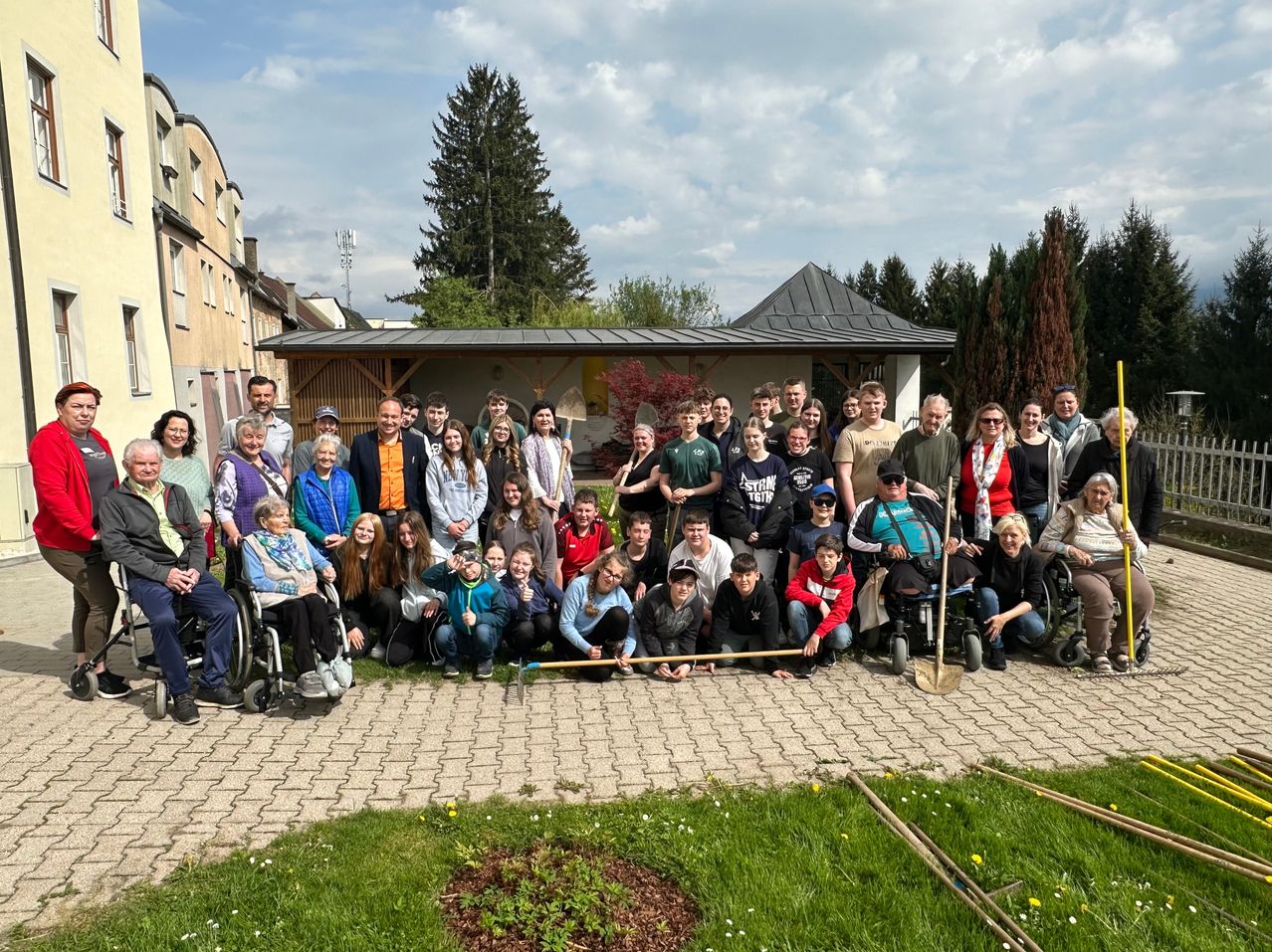 Eine Gruppe von Menschen in Rollstu?hlen und im Stehen posiert f?r ein Foto im Freien in einem Garten mit einem kleinen Baum in der Mitte.