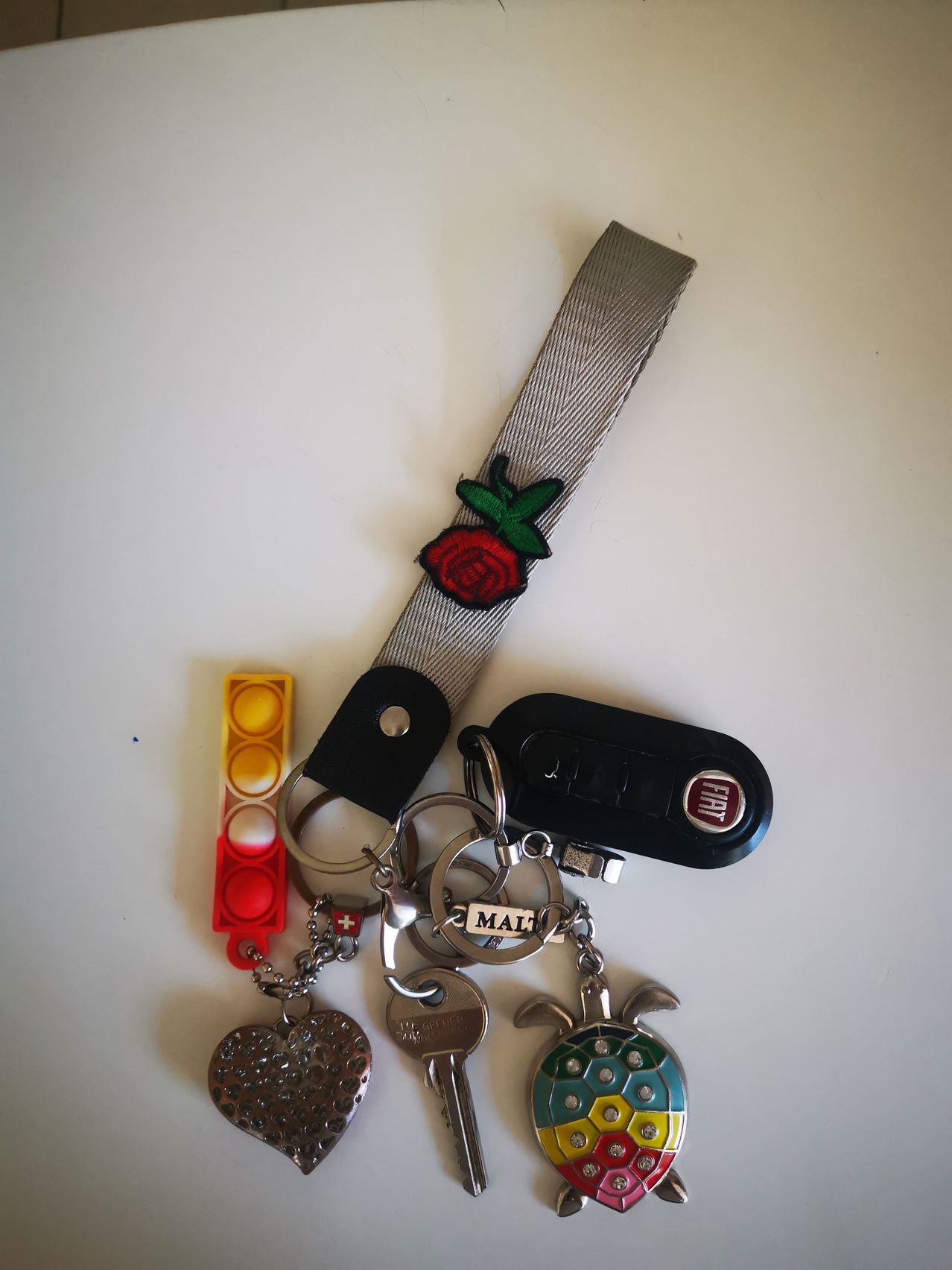 Ein Schlüsselanhänger mit verschiedenen Gegenständen enthält einen grauen Riemen, eine rote Rose, einen herzförmigen Schlüsselanhänger, einen Ampelschlüsselanhänger, einen Fiat-Schlüssel und einen Schildkröten-Schlüsselanhänger.