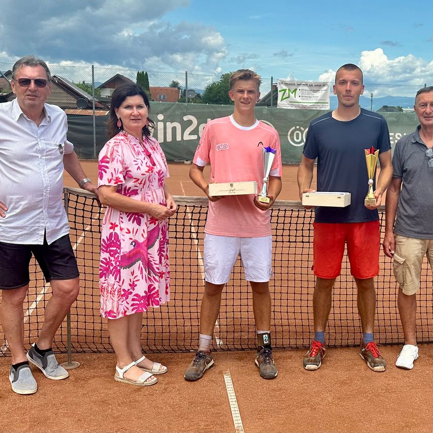 Fünf Personen stehen auf einem Tennisplatz. Drei von ihnen halten Pokale. Die beiden an den Seiten sind formell gekleidet, während die mittlere Person ein rosa Hemd trägt. Hinter ihnen steht ein Netz und ein Banner.