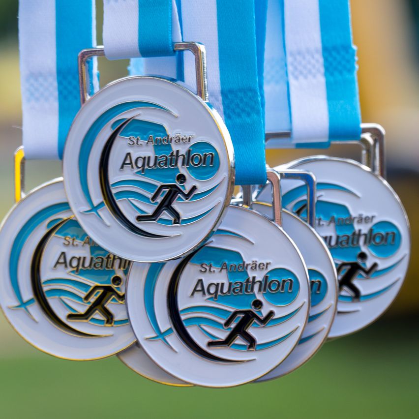 Eine Sammlung von Medaillen mit blauen und weißen Bändern. Die Medaillen haben ein Läufer- und Wellendesign mit dem Text 'St. Andräer Aquathlon'. Die Medaillen sind glänzend und metallisch.