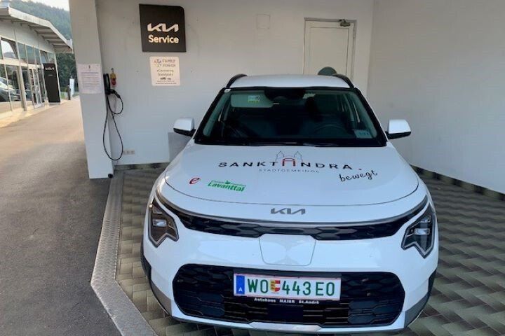 Ein weißer Wagen ist in einer Werkstatt geparkt. Er hat ein Nummernschild, das 'WO 443 EO' liest. Das Auto hat einen Slogan, der 'Santkadra' und 'bewegt' auf seiner Frontstoßstange liest. Hinter dem Auto ist eine Wand mit einem Schild, das 'Service' liest. Es gibt eine Steckdose, die an der Wand befestigt ist.