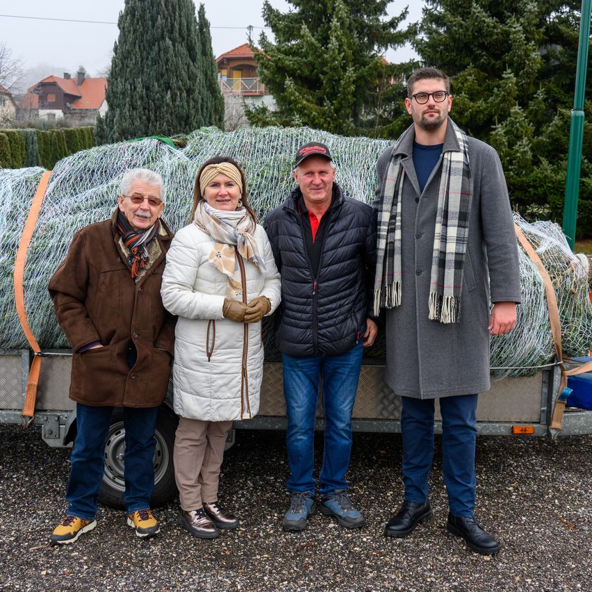 Vier Personen stehen vor einem mit Drahtgeflecht beladenen Anhänger, angezogen für kaltes Wetter. Dahinter sind Kiefern und Häuser zu sehen.