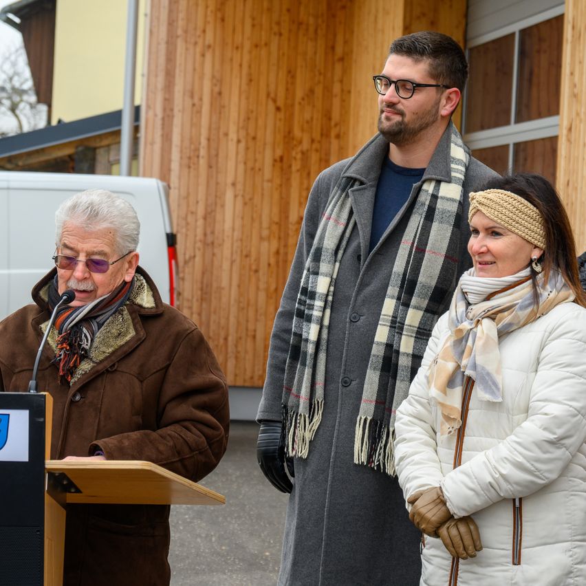 Ein älterer Mann spricht an einem Rednerpult, während zwei Personen in Winterkleidung hinter ihm stehen, in der Nähe eines Holzgebäudes und eines weißen Lieferwagens.