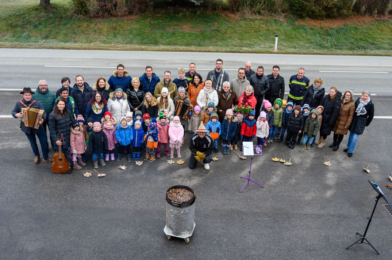 Eine große Gruppe von Menschen, einschließlich Kindern, versammelt sich um einen Topf auf einer Straße. Einige halten Musikinstrumente und es gibt Blätter auf dem Boden.