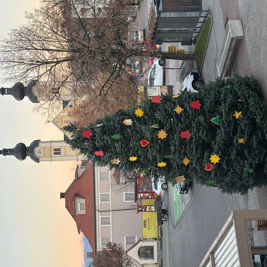 Luftaufnahme eines Weihnachtsbaums mit bunten Ornamenten auf einem Stadtplatz, Gebäuden und einem Uhrturm im Hintergrund.