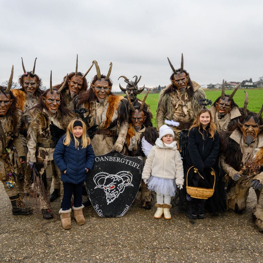 Eine Gruppe von Menschen in aufwendigen Kostümen, einschließlich Hörnermasken und pelzigem Kleidungsstücken, posiert für ein Foto. Zwei junge Mädchen in Engel- und Hexenkostümen stehen vor ihnen. Ein schwarzes Schild mit einem Widder und einigen Texten befindet sich in der Mitte.