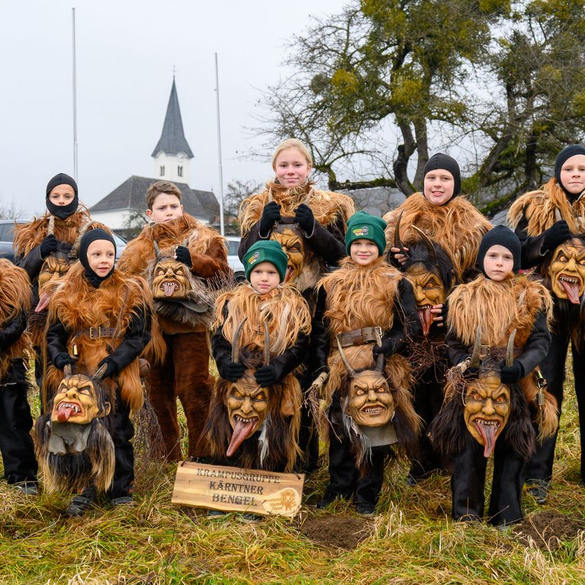 Eine Gruppe von Kindern in Krampus-Kostümen posiert für ein Foto vor einer Kirche mit einem Kirchturm. Sie halten Masken mit großen Mündern in den Händen.