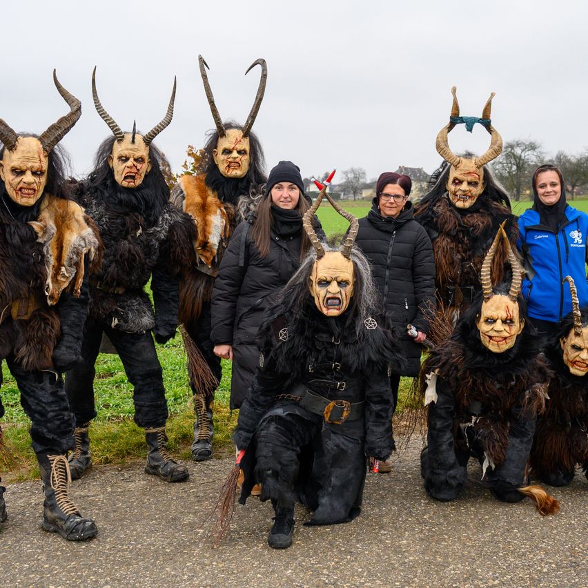 Eine Gruppe von Menschen in Hörnermasken posiert für ein Foto auf einer Straße, wobei zwei Frauen in Jacken in der Nähe stehen.