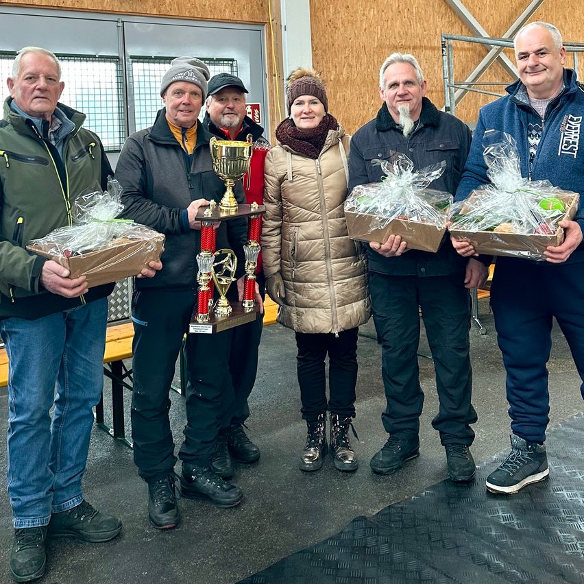Sechs Personen stehen zusammen und halten jeweils eine Trophäe und einen Geschenkkorb. Ein Mann hält eine goldene Trophäe mit roten Bändern. Die anderen tragen Geschenkkörbe, die in Plastik eingewickelt sind. Sie sind in Winterkleidung gekleidet. Hinter ihnen steht eine Holzbank und eine Wand.