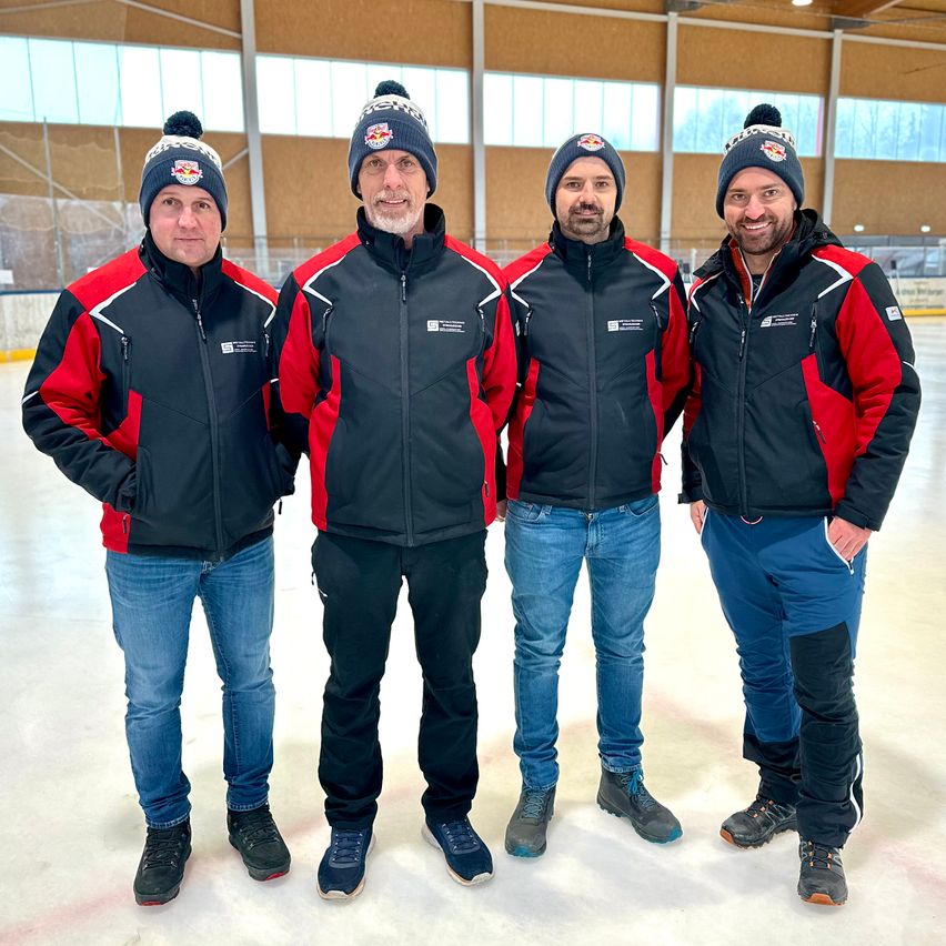 Vier Männer in Winterjacken und Mützen posieren für ein Foto auf einer Eisbahn. Sie lächeln und scheinen für ein Gruppenfoto zu posieren.
