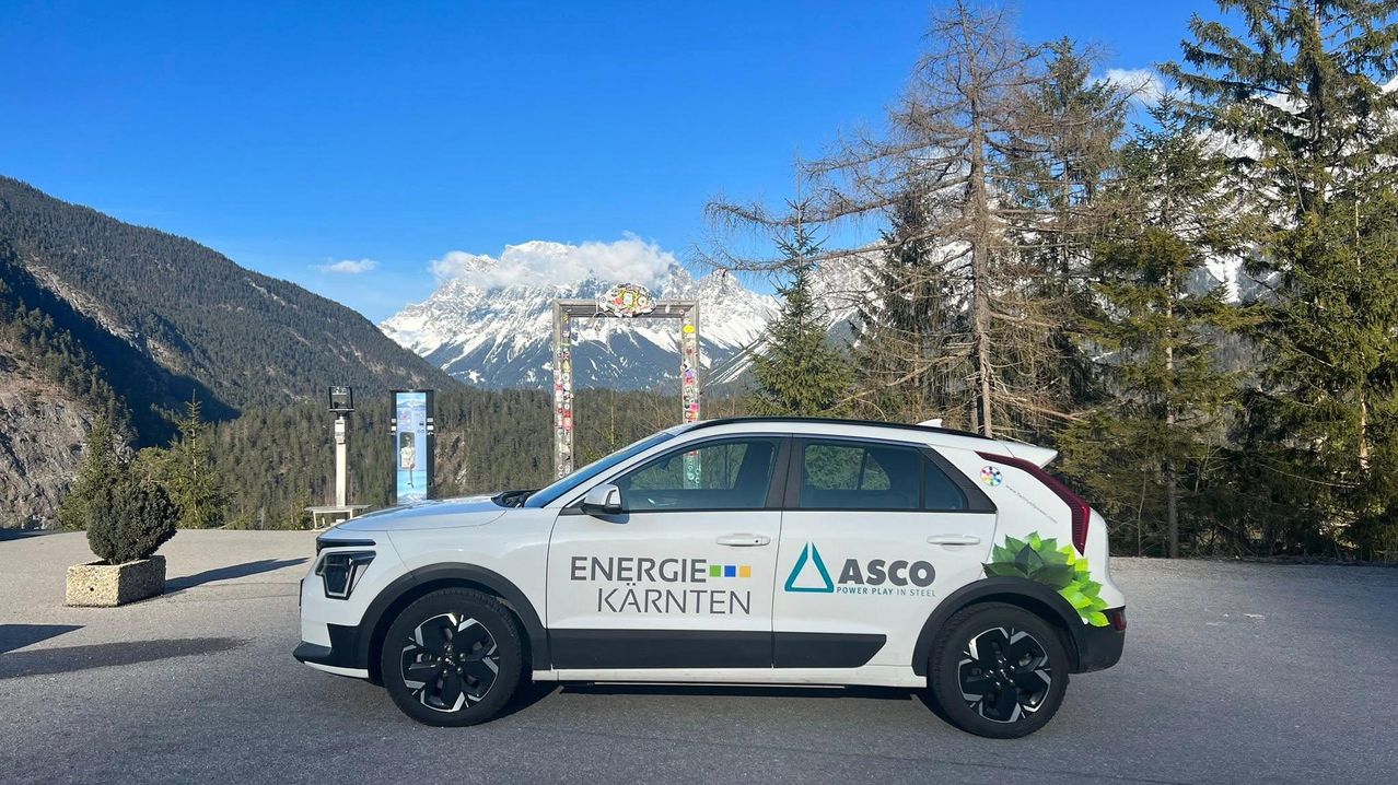 Ein weißer SUV mit 'Energie Kärnten' und 'ASCO Power Play in Steel' Logos ist in einem malerischen Gebiet mit schneebedeckten Bergen und grünen Bäumen geparkt.