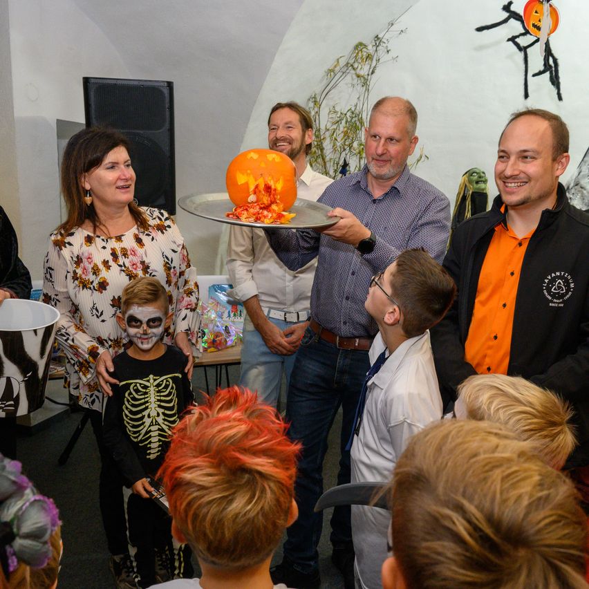 Mehrere Erwachsene und Kinder versammeln sich für eine Kürbisschnitzer-Veranstaltung. Ein Mann hält einen geschnitzten Kürbis auf einem Tablett. Ein Junge mit Skelett-Gesichtsbemalung steht neben einer Frau. Ein Mann hält einen Eimer.