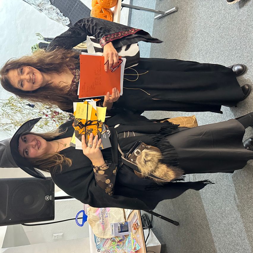 Zwei Frauen in Halloween-Kostümen, eine hält ein Buch und eine Tasche, während die andere ein Buch und einige Karten hält. Sie lächeln und posieren für ein Foto.