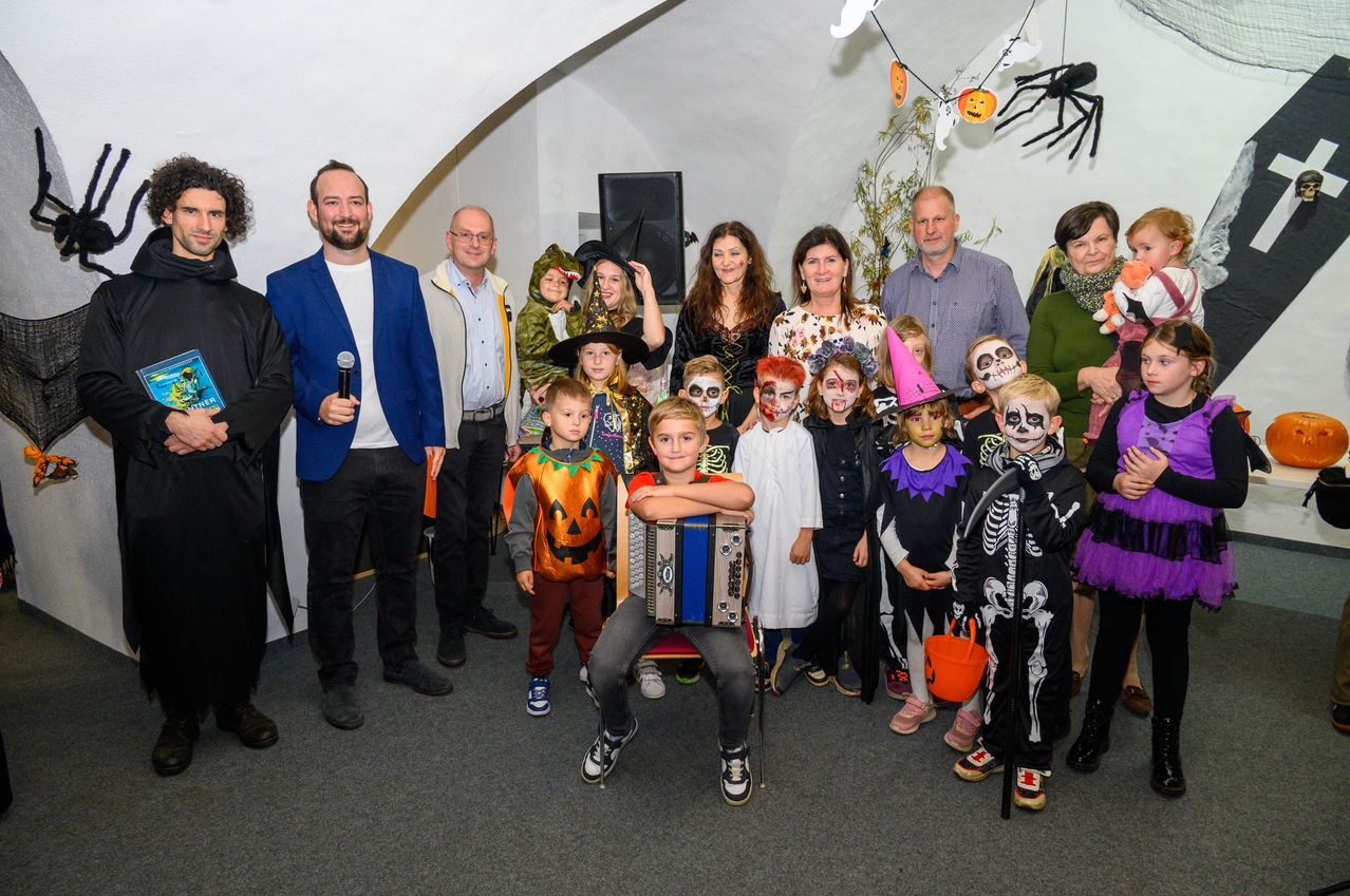 Eine Gruppe von Menschen, darunter Erwachsene und Kinder, posiert für ein Halloween-Foto. Einige sind in Kostümen gekleidet, und ein Junge spielt Akkordeon.