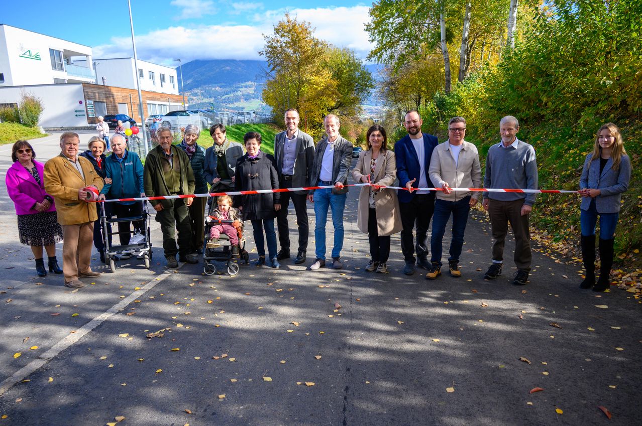 Eine Gruppe von Menschen steht auf einem Parkplatz und hält eine Schleife für eine Schleifenschneidezeremonie. Ein Baby in einem Kinderwagen ist anwesend. Bäume und Berge sind im Hintergrund.