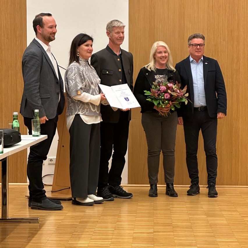 Fünf Personen stehen auf einer Bühne mit einer Holzwand im Hintergrund. Eine Frau hält ein Zertifikat und Blumen. Sie alle sehen glücklich aus und sind formell gekleidet.