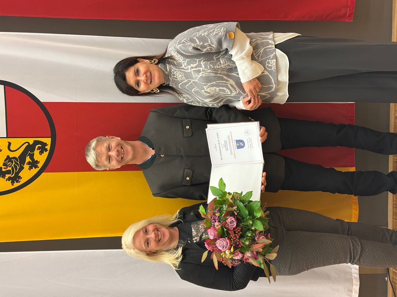 Drei Frauen posieren vor einem deutschen Flaggenhintergrund, eine hält ein Zertifikat, eine andere einen Blumenstrauß und die dritte liegt lächelnd auf dem Boden.