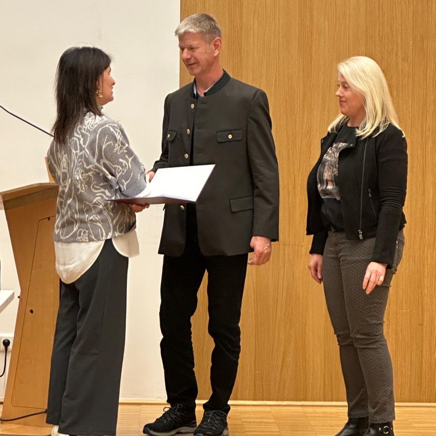 Drei Personen stehen in einem Raum zusammen. Eine Frau hält ein Papier und scheint es einem Mann zu präsentieren. Eine andere Frau steht neben ihnen. Sie alle lächeln.