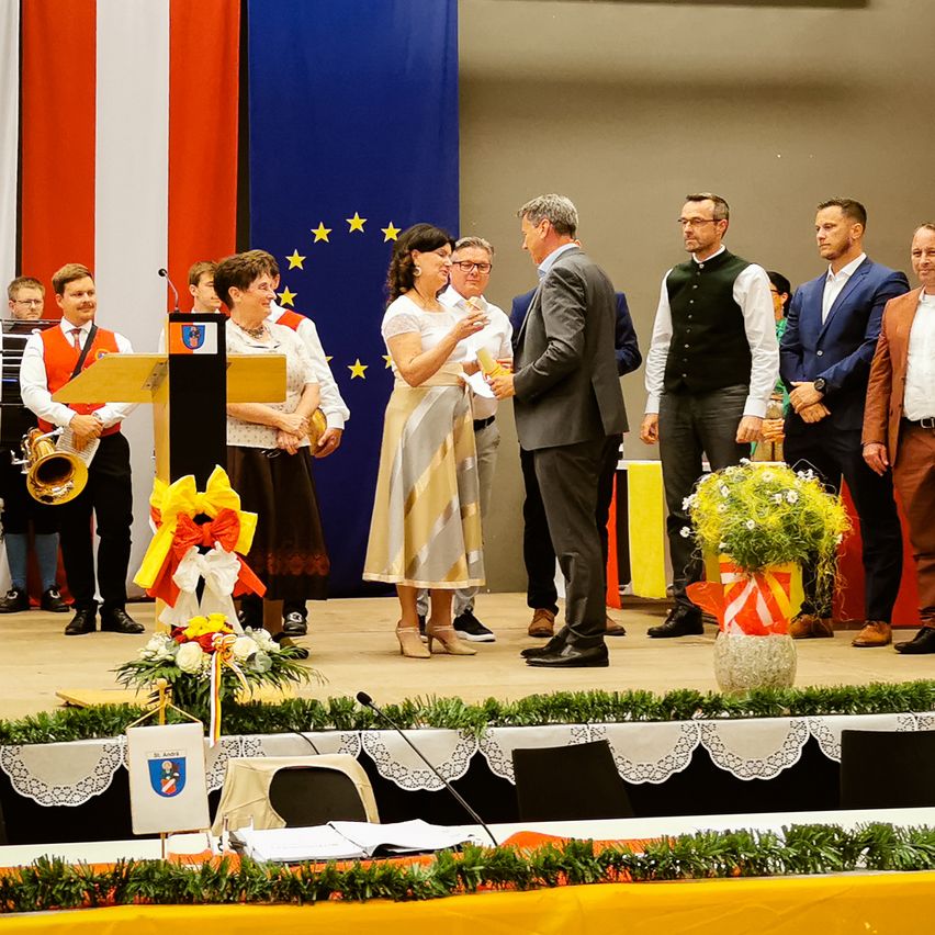 A group of people on a stage, one man shaking hands with another, a podium with flowers, and two men playing musical instruments.
