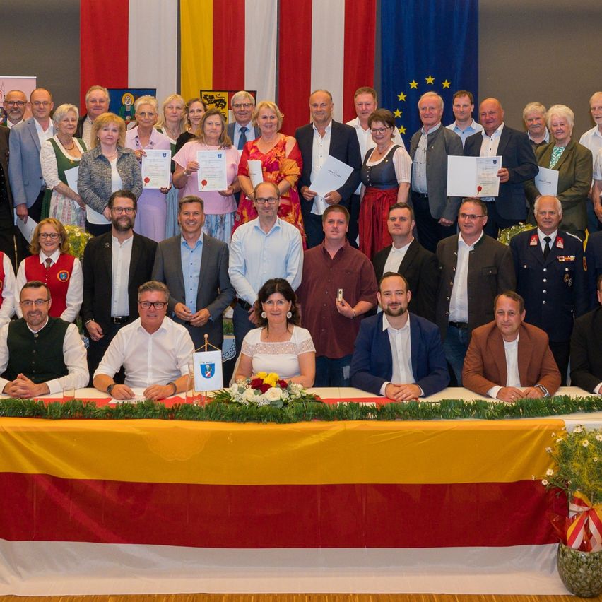 A group of people, including men and women, stand together for a picture. They hold certificates and some wear uniforms. A long table with flowers sits in the foreground.