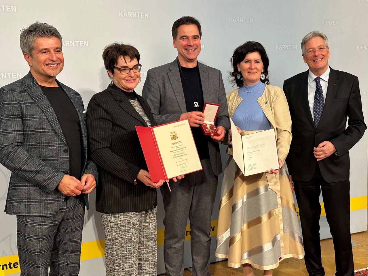 Fünf Menschen stehen in einer Reihe, halten Auszeichnungen und lächeln. Eine Frau links hält eine rote Auszeichnung, eine andere Frau rechts eine weiße. Dahinter befindet sich eine weiße Wand mit dem Text 'Kärnten'.