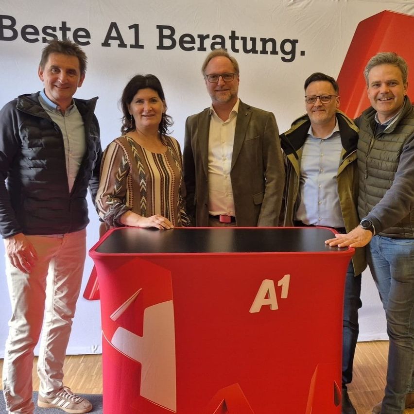 Fünf Personen stehen hinter einem roten A1-Schild, lächeln und posieren für ein Foto. Der Hintergrund zeigt ein weißes Banner mit dem Text 'Beste A1 Beratung.' Der Boden ist aus Holz.