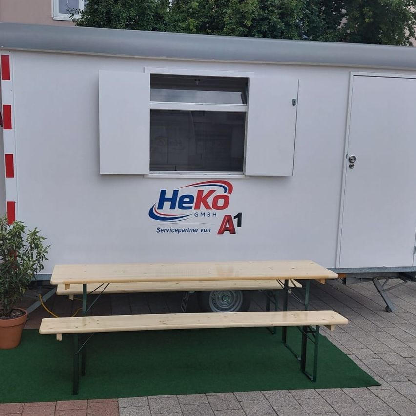 Ein weißer HeKo A1 Servicepartner-Anhänger mit offenem Fenster und einem Holztisch und Bänken davor.