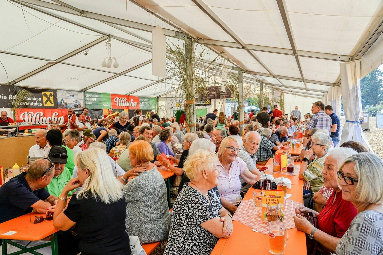 Eine große Gruppe älterer Menschen sitzt an langen Tischen in einem Zelt, einige unterhalten sich und andere genießen ihre Mahlzeiten. Die Tische sind mit karierten Tischdecken geschmückt, und es gibt Getränke und Essen auf den Tischen.