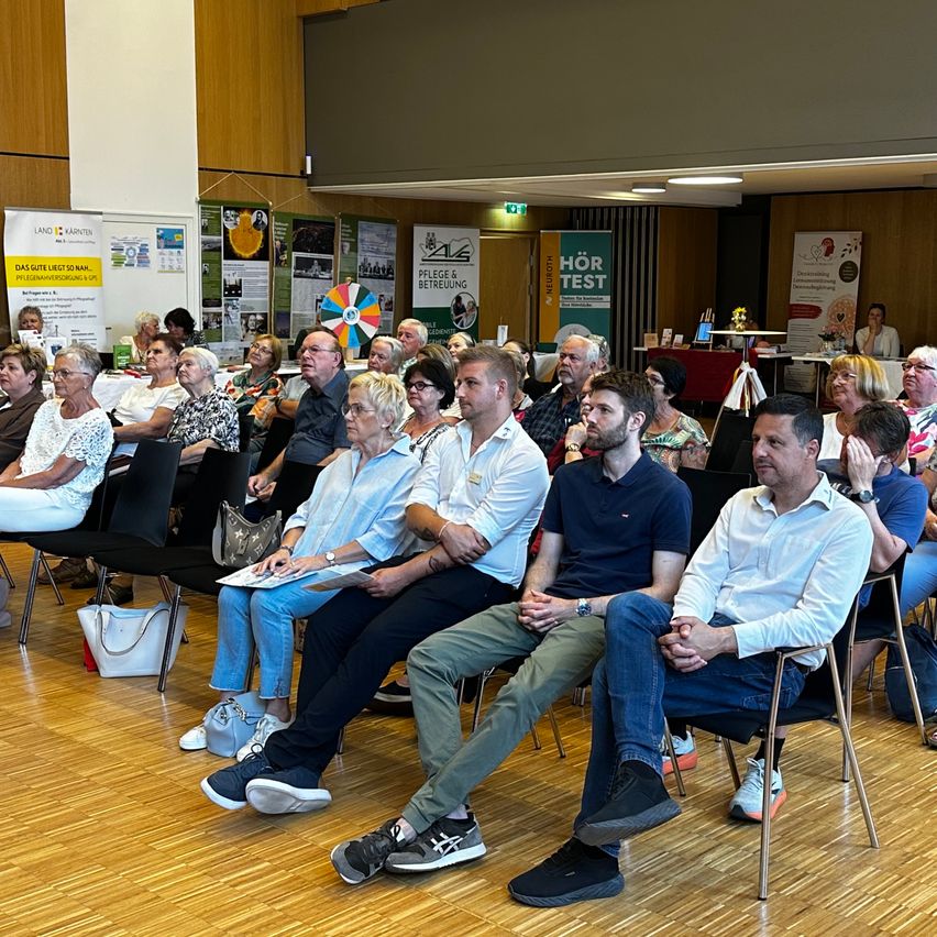 A group of people sit in chairs facing a stage, engaged in an event. Some hold papers or bags, and others watch attentively. Banners are displayed on the wall behind them.