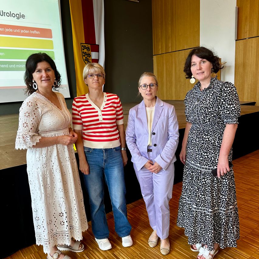 Four women stand on a stage, smiling and posing for a photo. Behind them, a projector screen displays 'Urologie'. The women wear various outfits, and the setting suggests a professional event.