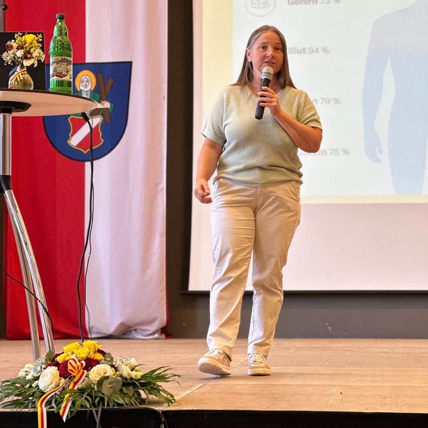 A woman speaks on stage with a microphone, flowers and a bottle in the background. Behind her is a projector screen displaying a silhouette with percentages.