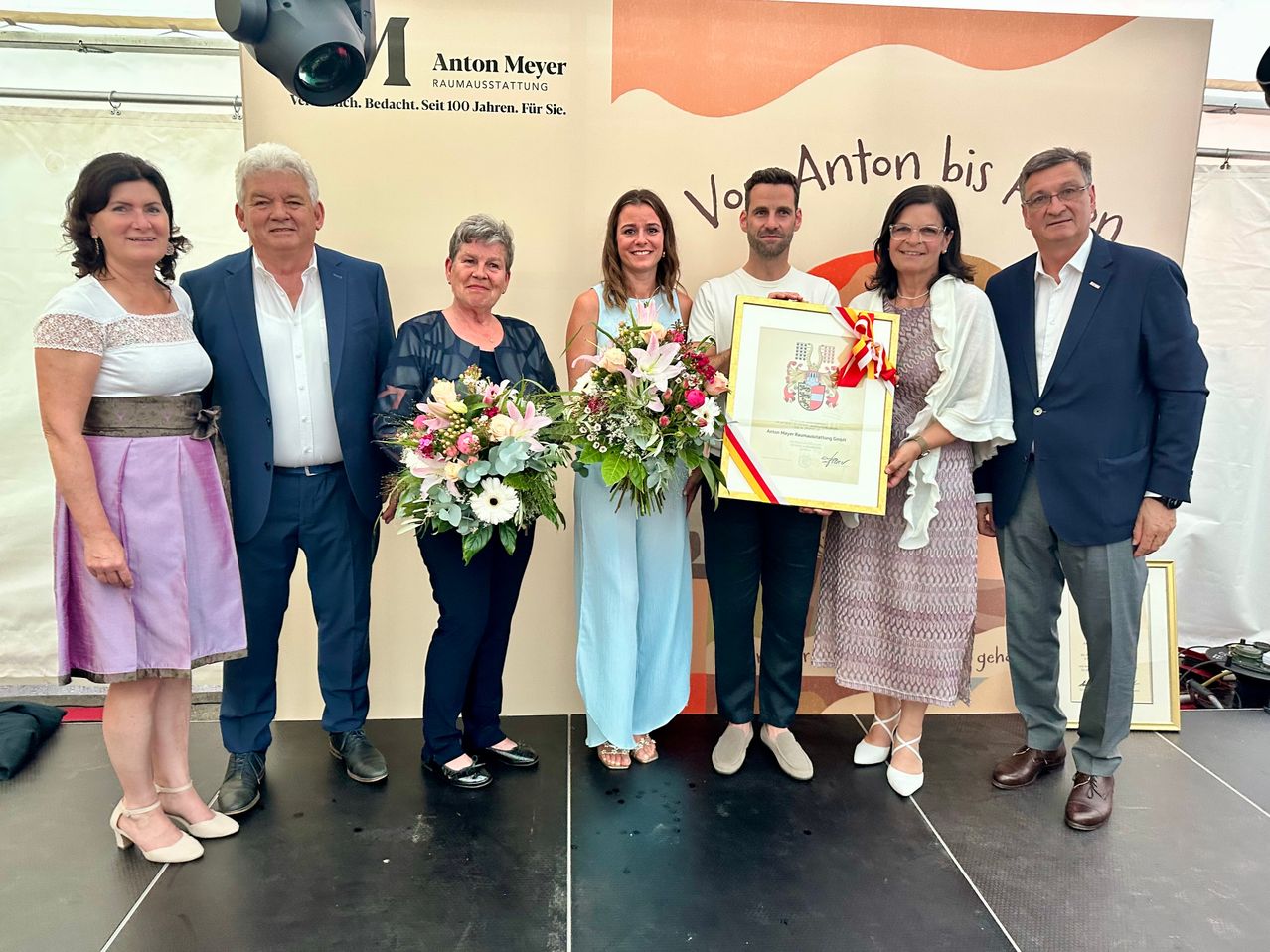 Eine Gruppe von Menschen posiert für ein Foto auf einer Bühne mit einem Zertifikat und Blumen in der Hand. Im Hintergrund ist ein Banner mit 'Anton Meyer' und 'Vom Anton bis' zu sehen.