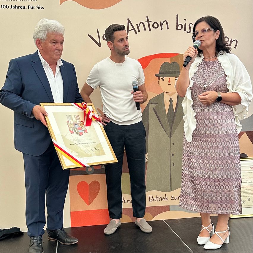 Drei Personen stehen auf einer Bühne und halten ein gerahmtes Zertifikat und ein Band. Ein Mann auf der linken Seite hält ein Mikrofon, während der Mann in der Mitte ein Zertifikat hält. Eine Frau auf der rechten Seite hält ein Mikrofon und spricht. Hinter ihnen befindet sich eine Wand mit einem Herz, einem Mann und einem Text.