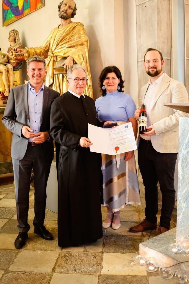 Vier Personen stehen lächelnd vor einer goldenen Statue. Der Mann in Schwarz hält ein Zertifikat, und der Mann in Weiß hält eine Flasche. Hinter ihnen steht eine Frau in einem blauen Kleid zwischen dem Mann mit Brille und dem Mann im Anzug.